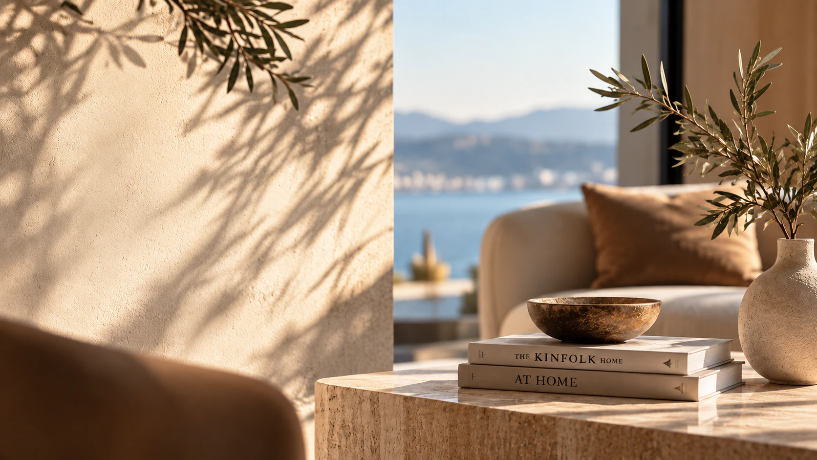 A sunlit living room: olive branches, a stone side table with books and a bowl, and a view over the water